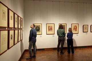 Drei Personen sind von hinten zu sehen, wie sie Kunstwerke an einer hellen Wand betrachten