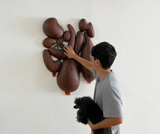 artist holding a black poodle, inspecting a wooden wall-mounted sculpture