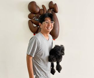 artist wearing yellow shorts and smiling to camera, holding a black poodle