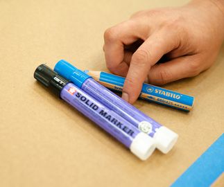 artist's picking up a chunky blue pencil, which is resting next to two marker pens on a brown work surface