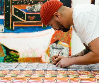 artist signing a stack of prints while wearing an orange beanie hat, a dark-toned painting is hanging on the wall behind him