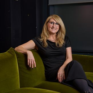 Anne Stagg, CEO of Digitas UK with long blonde hair and glasses sits relaxed on a green sofa, wearing a black outfit, with bookshelves in the background.