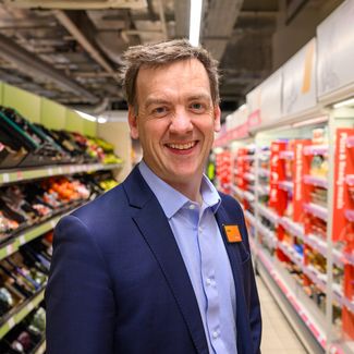 Mark Given, Sainsbury's marketing chief in a suit stands in a grocery store aisle stocked with fresh produce and packaged goods.