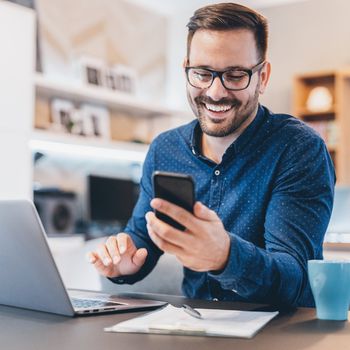 Smiling man with glasses using a smartphone at a desk, with an open laptop, notepad, and blue mug in a modern office setting.