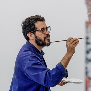 artist wearing blue shirt and glasses holding a long paintbrush in one hand and a paint palette in the other
