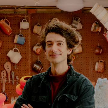 A close-up portrait of Maxwell Mustardo in his studio with hanging mugs on the wall behind him