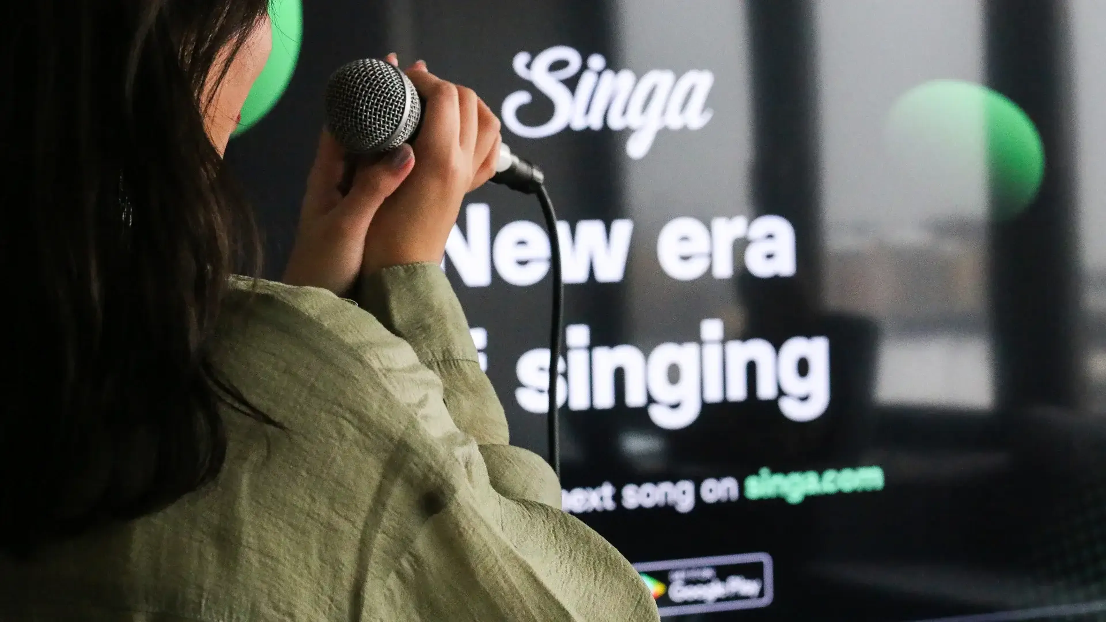 Person holding a microphone sings in front of a screen displaying "Singa: New era of singing."