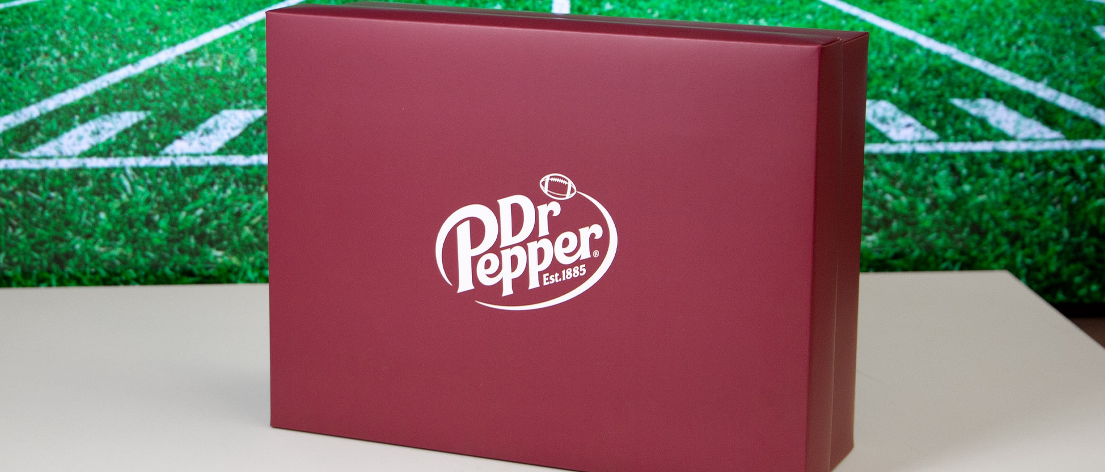 Rote Schachtel mit Dr Pepper Logo auf weißem Tisch und Foto von Football-Feld im Hintergrund