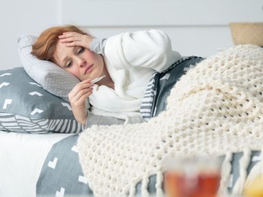 Eine Frau liegt im Bett, hält sich die linke Hand an die Stirn und hält in der rechten Hand ein Fieberthermometer.