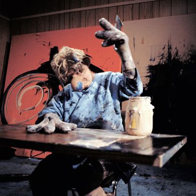 artist wearing a bulbous fake nose and gloves with large, sausage-like fingers – covered in paint