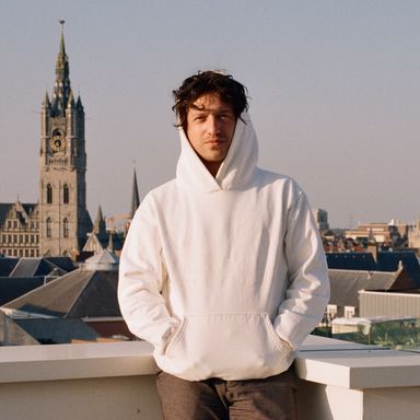 David Rudnick looking pensive on a rooftop, wearing a white hoodie with his hands in his front pocket