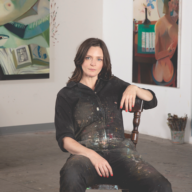 Danielle Orchard sitting in a chair in her studio with paint covered overalls on
