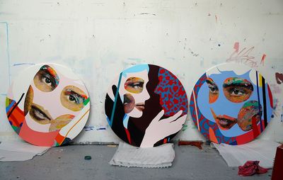 white wall with three circular tondo paintings propped against it of different faces depicted in different bold colours 