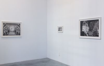 The corner of a gallery, with white walls and a concrete grey floor. There are two framed paintings on the wall to the right, and another left, all in black and white
