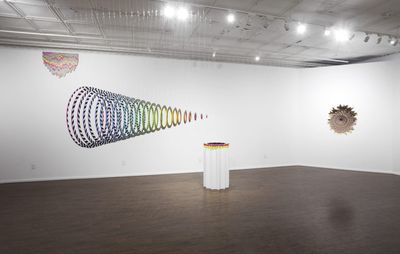 gallery view of spiralling coloured installation with several other artworks against white wall