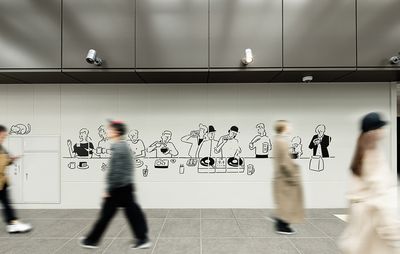 A white tiled wall on a street, decorated with line drawn images of people Djing, eating and drinking, by Yu Nagaba, while blurred people walk past