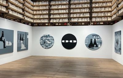 Installation view of white walls beneath shelves of books, where monochrome paintings are hung up