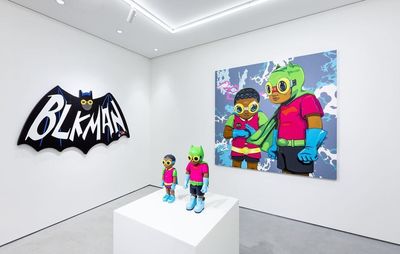installation view of a painting of cartoon superheroes alongside a bat-shaped artwork, with two small figurines on a white plinth