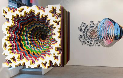 installation view of two kaleidoscopic sculptures suspended in mid-air using clear string with a patterned painting hung on the wall