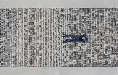 Ai Weiwei photographed from above, lying on his back with his arms behind his head amidst a neatly arranged grid of stones – grouped by size.