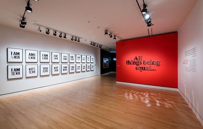 installation view of a red gallery wall introducing an exhibition title, opposite a white wall with twenty framed textual works of art