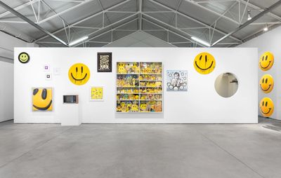 installation view of large freestanding white wall with yellow smiley faces hung all over it