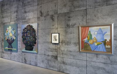 4 paintings against a star, concrete will in a gallery. One framed work, on the far right, includes a blue teapot on a table, against a sky blue background dotted with clouds.