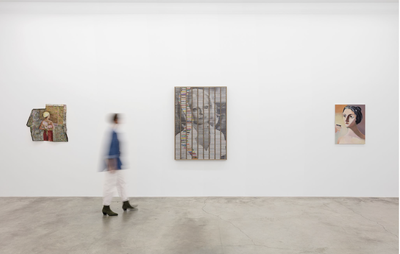 three paintings by various artists hanging on a white wall, where a blurred figure walks past