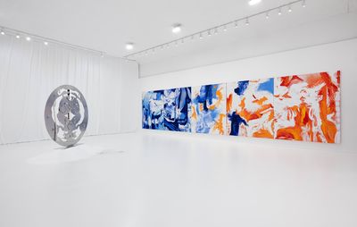 installation view of large white exhibition space with a free-standing metallic sculpture and a large horizontal painting made up of orange and blue tones