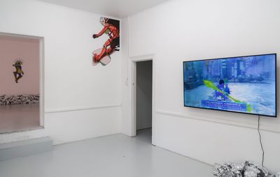installation view of gallery space with tv screen on one wall, a pile of silver foil below it, and a cartoon image above the doorway in the corner