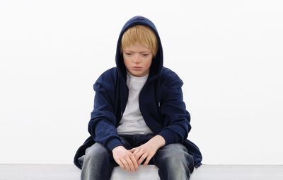 sculpture of a teenage boy with blond hair sat looking towards the ground with a blue hood over his head