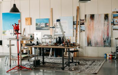 studio filled with various paintings on the walls and materials and brushes on a large work surface placed centrally
