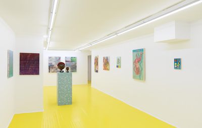 A white gallery space with a yellow floor. An array of small paintings hanging on the walls