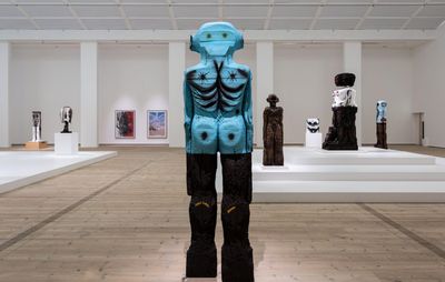 installation view of large blue figurative sculpture with more sculptures on plinths and paintings hung on walls in the background
