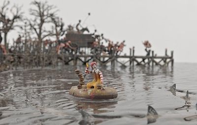 a clown floats on a small lifeboat on a body of water whilst figures battle in the background on a jetty
