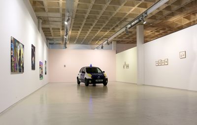 installation view of paintings in a white gallery space, with a car placed in the middle of the room