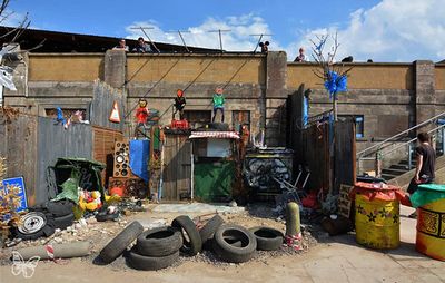 installation outside of a building with three figures stood on rooftops above a scene of junk objects, such as old car tyres, speaker systems, road signs, graffiti'd walls and debris