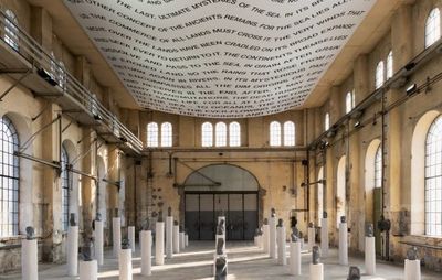 installation view of large exhibition space with writing covering the ceiling, and dozens of white plinths of varying heights with small sculptures placed atop of them