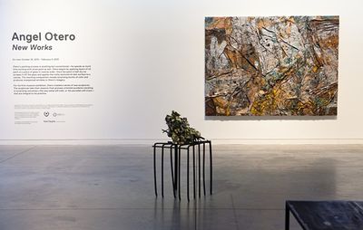 installation view of white wall with painting hung and exhibition title and description to the side of it, with a sculpture placed centrally on the floor