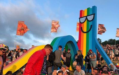 crowds of people stand and sit outdoors surrounding a large rainbow installation in the shape of a deckchair with sunglasses on and a smile 