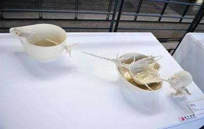 a white table with a white sculpture of a babies head blowing forward through a porcelain bowl and sculpted liquid shooting forwards into another porcelain bowl