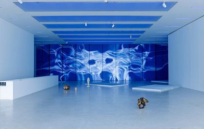 installation view of a large blue wall in a room with freestanding sculptures