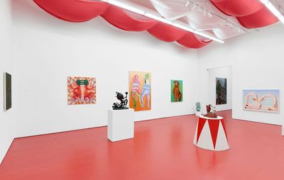 A photo of an art gallery with a red floor. The ceiling is lined with red and white material, and there are two artworks in the centre of the room, along with colourful art along the walls