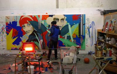 artist wearing black hat and blue coat working in his studio with heat lamp on and paint pots and cans on surfaces and on the floor, with a large painting on the wall in front of him