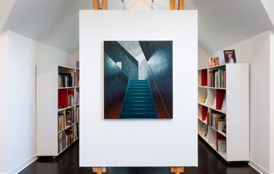 installation view of a painting hung on a white background placed on an easel with book shelves behind it