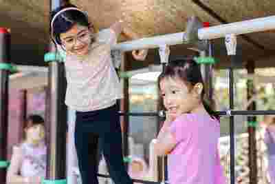 Two young girls playing on a climbing structure at a playground, one wearing glasses and the other in a pink shirt, both smiling.