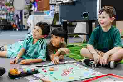 Three boys sitting on the floor, smiling and playing Monopoly, with game pieces and money spread out in front of them.