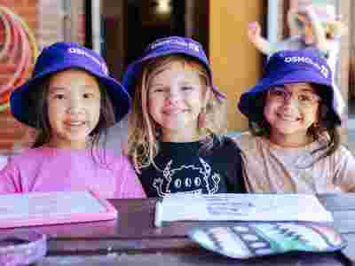 Three smiling children wearing purple OSHClub hats sit at a table with drawings. Another child is in the background, playing.