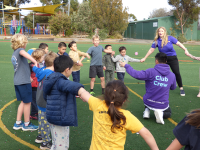 Children and two adults form a circle holding hands on a sports field, participating in a group activity. A playground is visible in the background.
