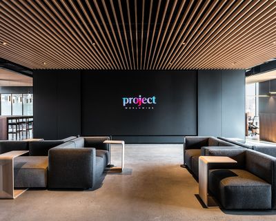 A large screen with the word "project" on display sits within a large black wall. Dark couches with wooden tables are placed around the room and the ceiling has vertical wooden panels decorating it.
